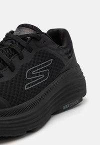 Zapatilla deportiva negra con parte superior de malla, suela gruesa de espuma y acentos grises. Presenta cierre de cordones y la marca "MAX CUSHION" en el lateral.