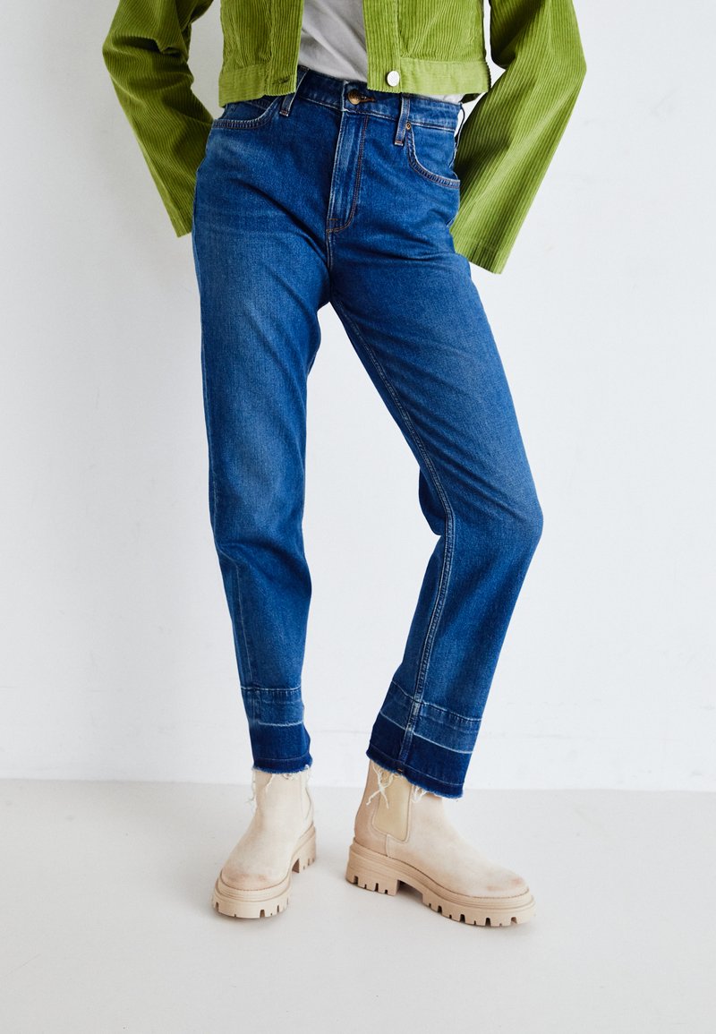 Jeans de mezclilla azul de cintura alta, pierna recta y dobladillos acampanados. Combinados con botas de tobillo beige y una chaqueta de pana verde.
