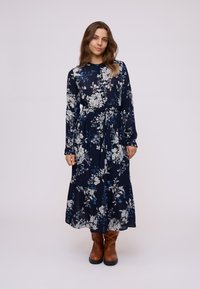 Robe maxi florale en bleu marine avec des manches longues, une taille nouée et un ourlet en tiers. Présente des motifs floraux bleu clair et blancs. Bottes marron.