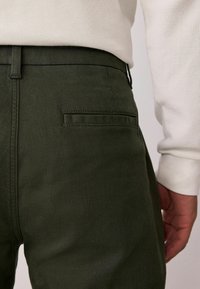 Dunkelgrüne Hose aus strukturiertem Stoff, mit zwei hinteren Taschen und einem nahtlosen Bund. Der Hintergrund ist neutral.