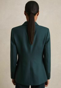 Tumši zaļš pielāgots bleizeris ar strukturētu piegriezumu, ar izgrieztiem atloka galiem un pogātām aprocēm, kas izceļ gludu audumu un skaidru siluetu.