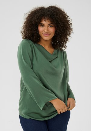 Haut long à manches vert, texturé, avec un col drapé et une coupe décontractée, présenté sur un jean bleu foncé et assorti à un fond neutre.