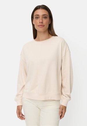 Frau mit glatten braunen Haaren, die einen hellbeigen Langarmpullover und weiße Hose trägt, steht vor einem schlichten weißen Hintergrund.