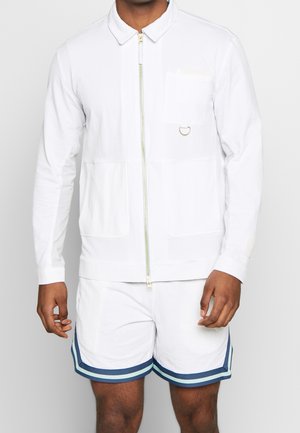 Mann trägt weiße Zip-up-Jacke mit Brusttasche und D-Ring, kombiniert mit weißen Shorts mit blauen und hellblauen gestreiften Saumkanten.