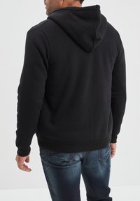 Sudadera negra con tejido texturizado, que presenta una capucha y puños de canalé, vista desde la parte trasera, combinada con jeans azul desgastados.