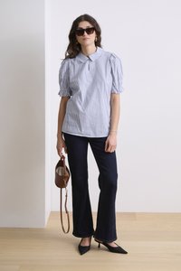 Blusa a righe blu e bianche con colletto e maniche a sbuffo, abbinata a jeans svasati scuri e tacchi a punta neri. Borsa marrone con tracolla.
