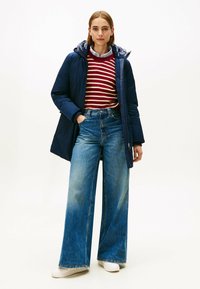 Giacca puffer blu navy con cappuccio, maglione a righe bordeaux e crema, jeans a vita alta e gamba larga, e sneakers bianche. Tessuto strutturato e vestibilità rilassata.
