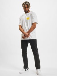 Witte katoenen t-shirt met een geel Wu-Tang Clan-logo, gecombineerd met zwarte jeans en witte Nike sneakers met zwarte accenten.