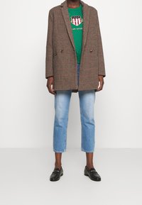Bruine geruit oversized blazer met geknipte revers, dubbelzijdige knopen, gedragen over een groene graphic sweatshirt en lichtblauwe cropped jeans.
