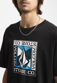 Sort T-shirt med korte ærmer, der har en stor, fed grafik i blå og hvid, der viser et logo og geometriske former. Sølv kæde halskæde.