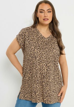 Kvinde med langt brunt hår iført en kortærmet V-hals leopardmønstret top og blå jeans, stående med den ene hånd på hoften.