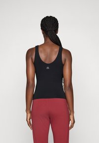 Schwarzes Tanktop mit einem runden Rückendesign, aus dehnbarem Material gefertigt. Hat ein kleines Logo in der Mitte des Rückens. Kombiniert mit dunkelroten Hosen.