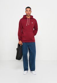 Maroon hoodie with drawstring, front pocket, and small logo. Worn with loose blue jeans and white sneakers, holding a black bag.