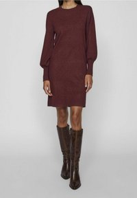 Tmavě červené pletené šaty s dlouhými puff rukávy, kulatým výstřihem a bočními rozparky, v kombinaci s vysokými hnědými koženými botami s přiléhavým designem.