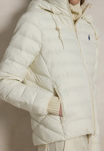 Persoon draagt een crème doorgestikte donzen jas met ritszakken en een capuchon, daarover een geribde crème coltrui.