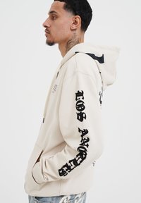 Felpa beige con grafiche nere sulle maniche e sul petto. Presenta un cappuccio con coulisse e una tasca a marsupio con una texture morbida.