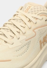 Béžová sportovní obuv s texturovaným síťovaným svrškem, tenkými tkaničkami a logem. Má polstrovanou podrážku a vzorovanou výzdobu na boku.