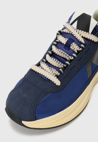 Navy blue sneaker with a textured upper, featuring beige laces, a padded collar, and a contrasting cream midsole. Rubber outsole with grip patterns.