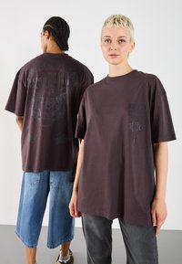 Mørkebrun oversized t-shirt med grafiske prints på front og bagside, lavet af blødt bomuldsstof. Casual stil med nedsatte skuldre og afslappet pasform.