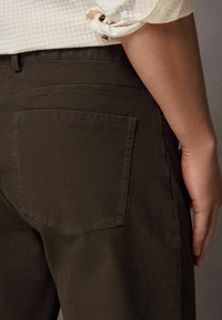 Bruine broek van gestructureerde stof, met een achterzak en een rechte snit, gecombineerd met een lichtgekleurde shirt met opgerolde mouwen.