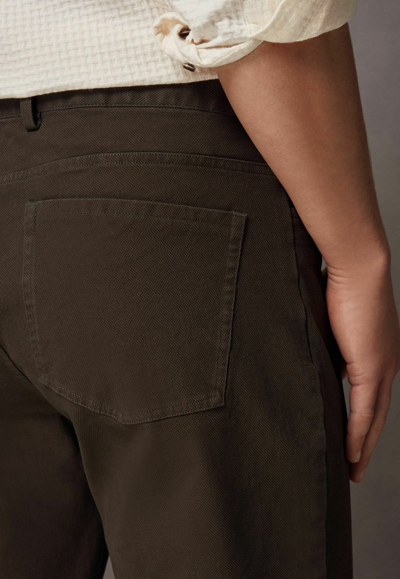 Bruine broek van gestructureerde stof, met een achterzak en een rechte snit, gecombineerd met een lichtgekleurde shirt met opgerolde mouwen.
