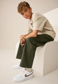 Béžová textúrovaná košeľa s krátkym rukávom, olivovo zelené široké nohavice a biele tenisky s hnedými detailmi. Dieťa sedí na zakrivenom svetlom povrchu.