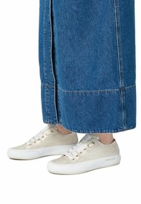 Sneakers med en strukturerad krämfärgad ovandel, vit gummisula och snörningsdesign. Bärs med en lång jeanskjol i blått, där endast fötterna syns.