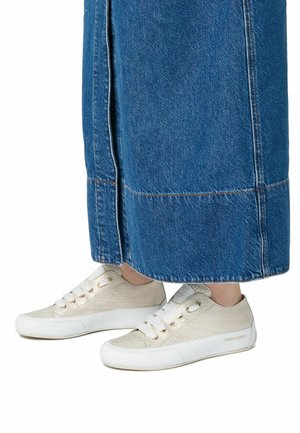 Sneaker con parte superiore in crema texture, suola in gomma bianca e design con lacci. Indossati con una lunga gonna in denim blu, mostrando solo i piedi.