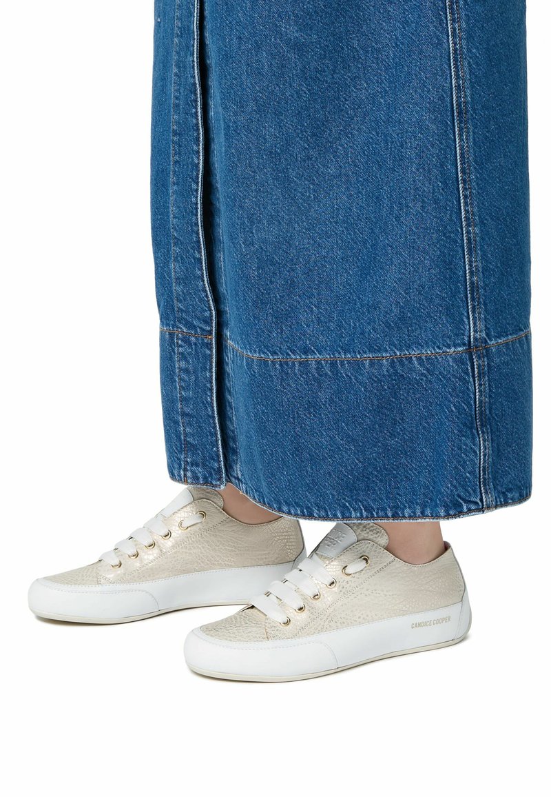 Sneakers med en strukturerad krämfärgad ovandel, vit gummisula och snörningsdesign. Bärs med en lång jeanskjol i blått, där endast fötterna syns.
