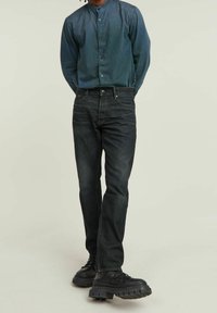 Jeans de mezclilla en un tono oscuro con un corte recto, acompañados de una camisa de botones índigo. Unos zapatos de suela gruesa negra completan el conjunto.