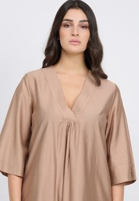 Top beige de cuello plegado hecho de una tela suave, con mangas de tres cuartos y un corte holgado, que cuenta con un sutil fruncido en la parte delantera.