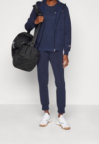 Person, der einen navyblauen Trainingsanzug und weiße Sneaker trägt und eine schwarze Sporttasche über der Schulter hält, vor einem einfarbigen Hintergrund.