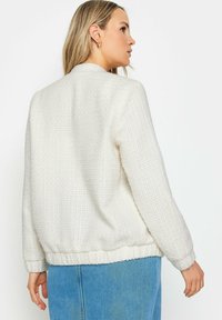 Maglione bianco testurizzato con scollatura a girocollo e maniche lunghe, caratterizzato da orlo e polsini a coste; indossato sopra una gonna in denim blu.