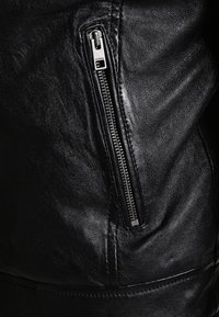 Veste en cuir noir avec une texture lisse, dotée d'une poche zippée en argent sur le côté gauche et de détails de couture le long des coutures.