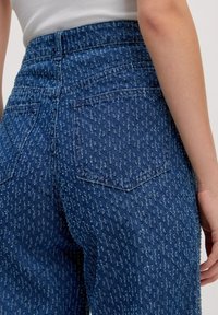 Hoch geschnittene blaue Jeans aus Denim mit einem strukturierten Muster. Zu den Merkmalen gehören aufgesetzte Taschen und ein gerades Beinprofil.