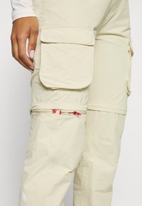 Beige arbetspants med flera stora fickor, dragkedjor med röda detaljer och en avslappnad passform. Hållbart material och mjuk textur.