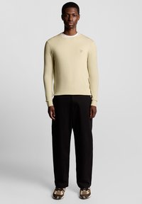 Suéter amarelo claro de mangas compridas com punhos canelados, combinado com calças pretas e sapatos castanhos com padrão. Design simples, ajustado, com detalhe de logótipo.