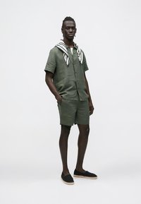 Camisa de linho verde-oliva de manga curta e calções combinando, com um lenço preto e branco com padrão pendurado nos ombros, e espadrilhas pretas.