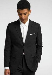 Chaqueta de traje negra con una tela texturizada, presenta dos botones, solapas de muesca y bolsillos laterales. Llevada sobre una camisa blanca de botones.