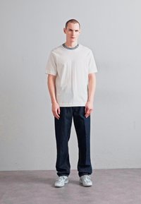 Tricou alb din bumbac cu guler gri, asortat cu pantaloni din denim de culoare bleumarin. Modelul stă într-un cadru minimalist, cu un fundal neutru.