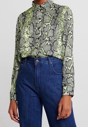 Osoba ubrana w zieloną i czarną koszulę na guziki z długim rękawem o wzorze węża, wsuniętą do wysokich, ciemnoniebieskich dżinsów.