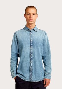 Koszula dżinsowa w jasnoniebieskim kolorze, z zapięciem na guziki z przodu, kołnierzykiem, długimi rękawami oraz kontrastującymi przeszyciami wzdłuż szwów.