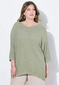 Maglione oversize verde chiaro con un design in maglia testurizzata, scollo rotondo e maniche a tre quarti. Abbinato a pantaloni di colore chiaro.