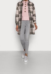 Personne portant un jean skinny gris, un pull rose clair avec de gros boutons, un long manteau à carreaux gris et rose, et des baskets gris, marchant vers l'avant.
