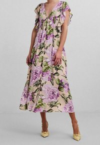 Robe maxi florale avec un décolleté en V, des manches courtes volantes, un fond beige clair, et de grands imprimés de fleurs violettes ; assortie à des chaussures jaunes à talons pointus.