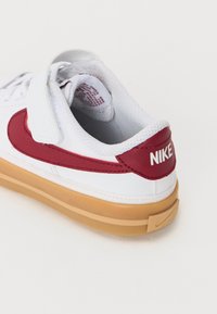 Beli Nike superge z rdečimi detajli, teksturirano zgornjim delom, Velcro trakom in rjavim gumijastim podplatom, ki ima Nike logo na peti.