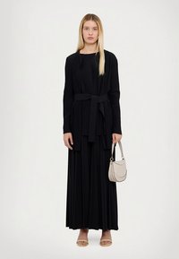 Melna plīsēta gara kleita ar saskaņotu jostu, garām piedurknēm un apaļu izgriezumu; komplektā ar bēša krāsas rokassomu un gaismas toņa sandales.