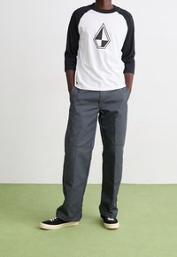 Langærmet, hvid baseball T-shirt med sorte ærmer, der har et diamantlogo. Grå, løstsiddende bukser med en glat tekstur og sidelommer. Sorte sneakers.