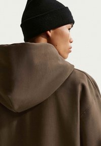 Persona joven con sudadera con capucha marrón y gorro de punto negro, de perfil hacia la derecha contra un fondo claro y liso.