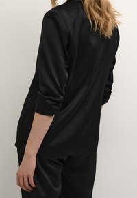 Musta bleiseri, jossa on sileä rakenne, pitkät hihat ja hieman muotoiltu design. Siinä on sivutasku, ja se on yhdistetty matching mustiin housuihin.